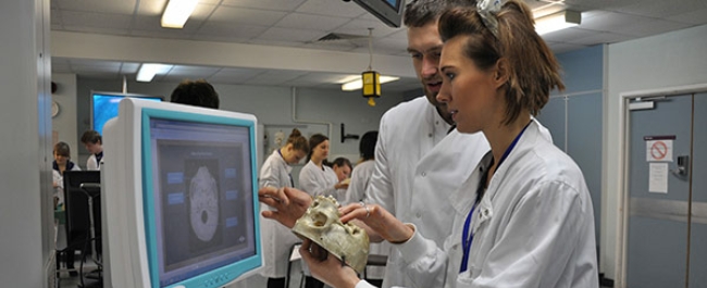 Two anatomy students in lab coats are studying a scan of a skull on a monitor. One of the students is holding part of a human skull.